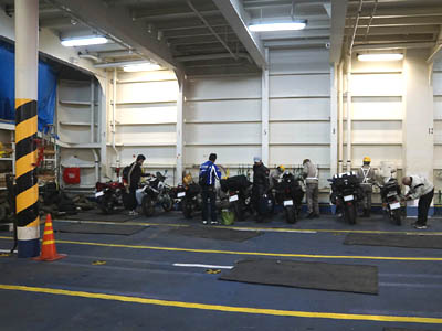 太平洋フェリー「いしかり」に乗船して車両甲板に駐車したバイクを固定している甲板員と荷降ろし中のライダー